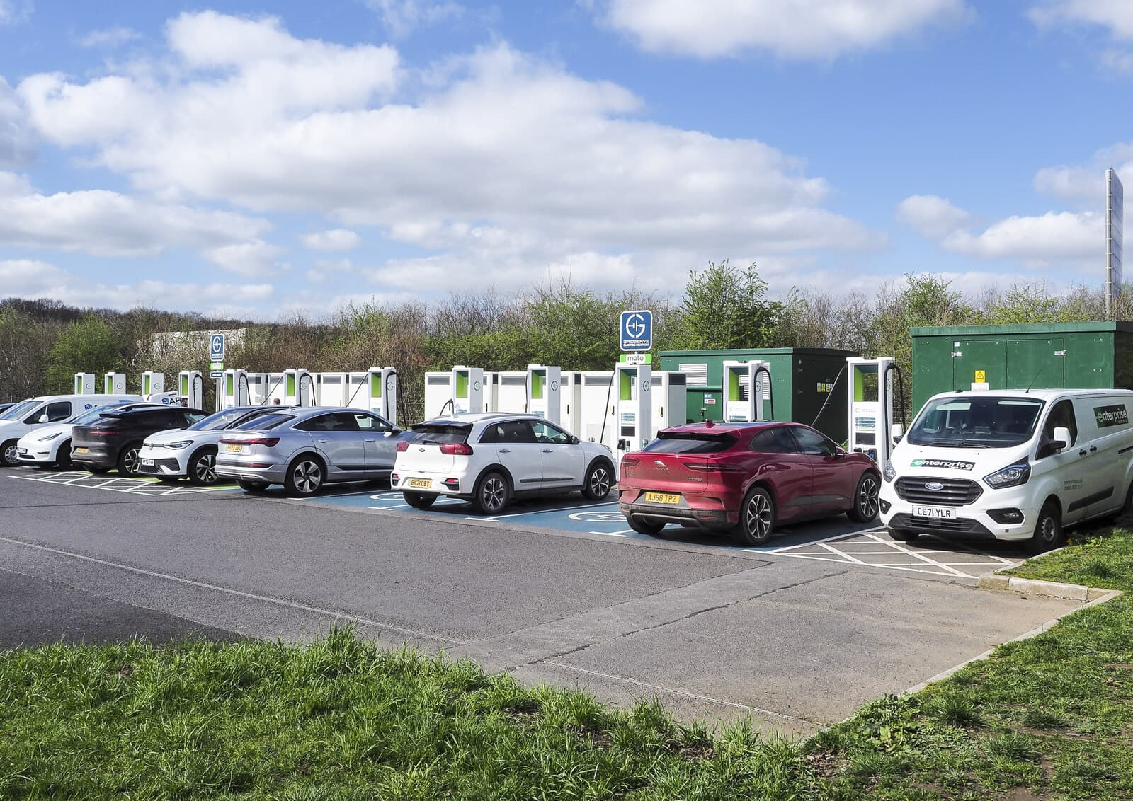 A row of rapid electric vehicle chargers in a UK car park, with several cars and a van plugged in.